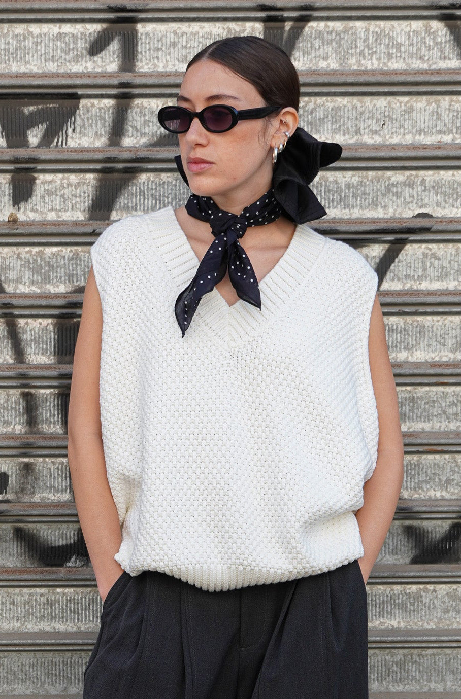Model wearing a white knitted vest and grey pants with a black polka dots scarf