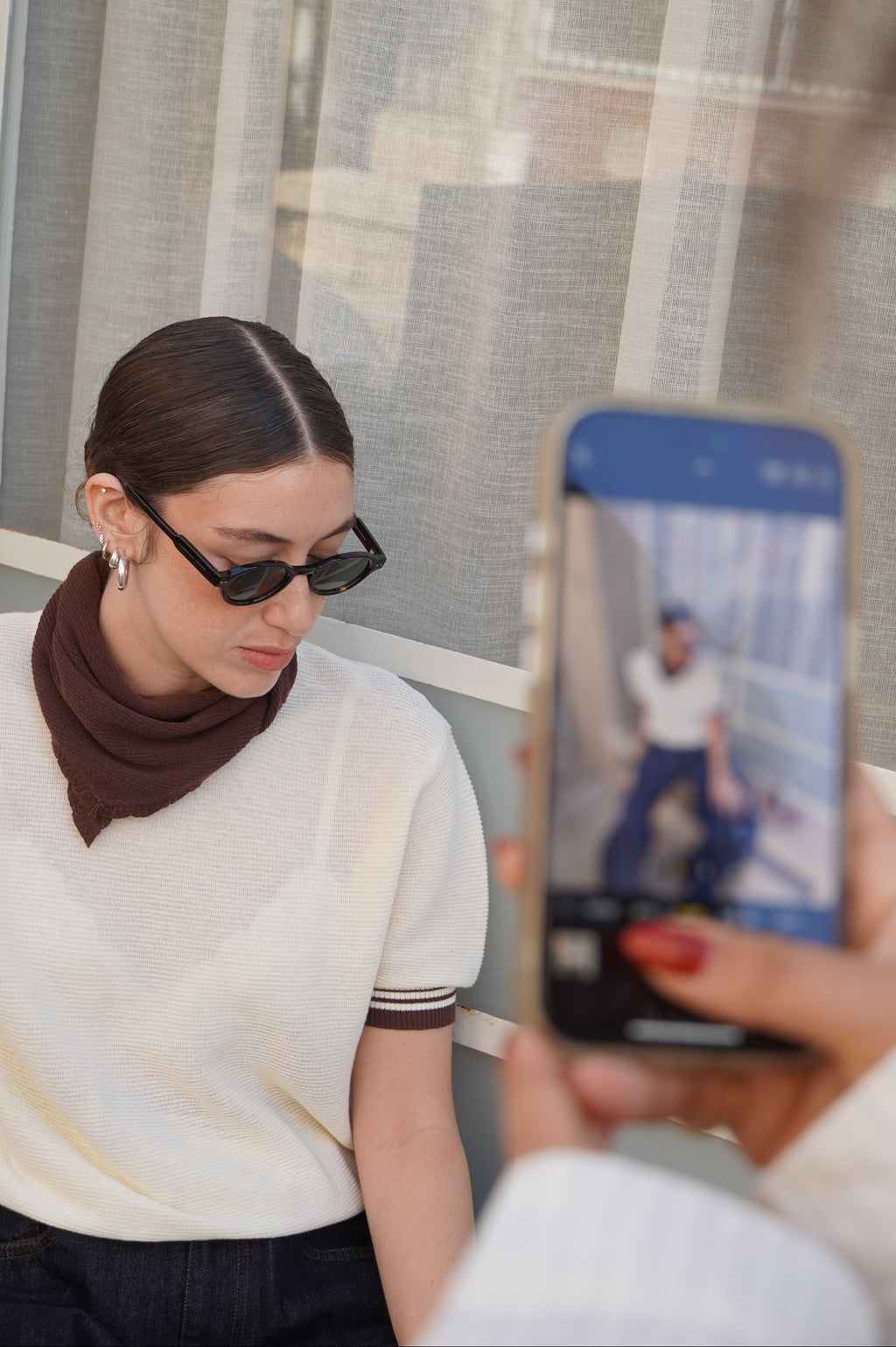 Person taking a photo of a model wearing a white shirt with brown sleeves, and a brown knitted scarf